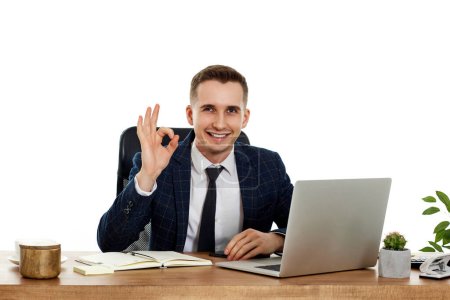 Téléchargez les photos : Jeune homme d'affaires heureux assis au bureau travaillant sur ordinateur portable PC et montrant geste Ok - en image libre de droit