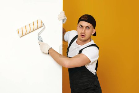 Foto de Hombre sorprendido en el trabajo de ropa de trabajo con rodillo de pintura, pintura de pared, banner web y espacio de copia - Imagen libre de derechos