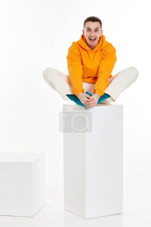 Photo for Full length photo of young man in orange sweatshirt sitting on white cube on white background. - Royalty Free Image
