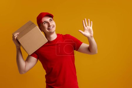 Téléchargez les photos : Heureux livreur employé en bonnet rouge, t-shirt rouge tenant boîte en carton isolé sur fond jaune - en image libre de droit