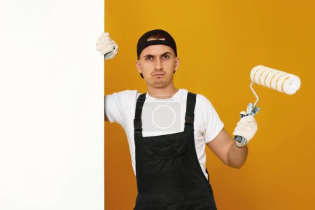 Photo for Frowning man in working clothes work with paint roller, wall painting, web banner and copy space - Royalty Free Image