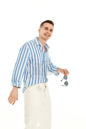 Foto de Joven guapo positivo en gafas de sol y camisa de rayas azules aislados sobre fondo blanco. Moda de verano - Imagen libre de derechos