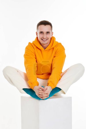 Photo for Young toothy guy in orange sweatshirt sitting on white cube on white background. Full length - Royalty Free Image