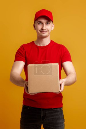Photo for Delivery man employee in red cap, red t-shirt holding cardboard box on yellow background. focus on box - Royalty Free Image