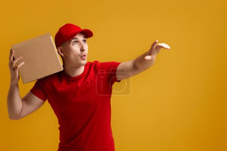 Photo for Delivery man employee in red cap, red t-shirt holding cardboard box and pointing finger at the side isolated on yellow background - Royalty Free Image