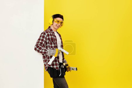 Foto de Guiño obrero hombre o constructor en casco de construcción cerca de bandera blanca sobre fondo amarillo. edificio carpintería profesión - Imagen libre de derechos