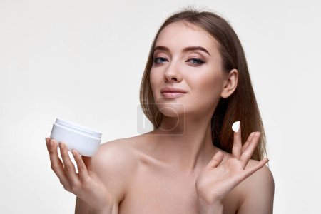 Foto de Hermosa chica sonriente aplicando crema para el cuidado de la piel en su cara y sosteniendo frasco de producto cosmético sobre fondo blanco - Imagen libre de derechos