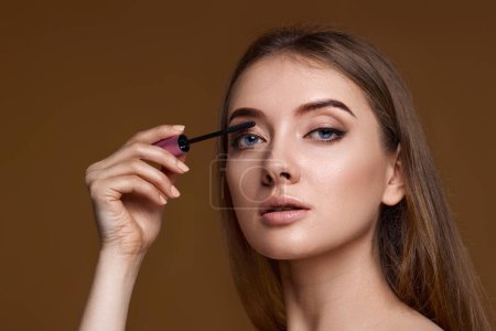 Photo for Beautiful woman applying black mascara on long thick eyelashes with brush on beige background - Royalty Free Image