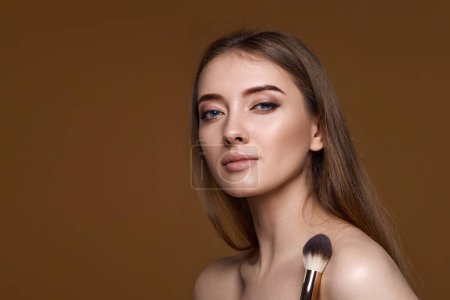 Téléchargez les photos : Jeune femme appliquant rougeur sur les joues avec pinceau de maquillage sur fond beige. espace de copie - en image libre de droit