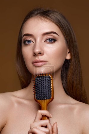 Téléchargez les photos : Belle femme brune montrant une brosse en bois et recommandant un produit de soins capillaires pour des cheveux longs et sains isolés sur fond beige - en image libre de droit