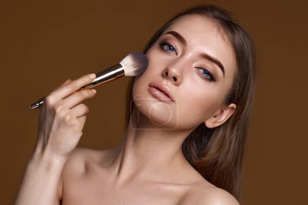 Téléchargez les photos : Jeune femme appliquant rougeur sur les joues avec pinceau de maquillage sur fond beige - en image libre de droit