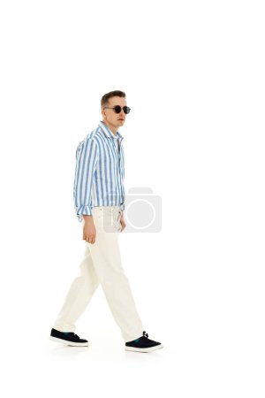 Téléchargez les photos : Souriant homme heureux confiant dans des lunettes de soleil et chemise à rayures bleues debout isolé sur fond blanc. sur toute la longueur. Mode d'été - en image libre de droit