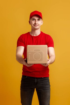 Photo for Delivery man employee in red cap, red t-shirt holding cardboard box isolated on yellow background - Royalty Free Image