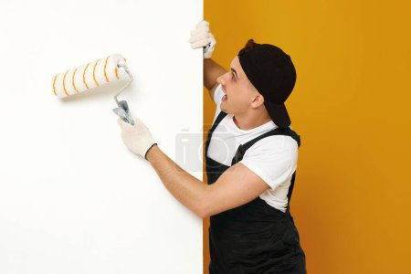 Téléchargez les photos : Ouvrier bricoleur en vêtements de travail peignant des murs blancs dans la pièce. espace de copie pour le texte - en image libre de droit