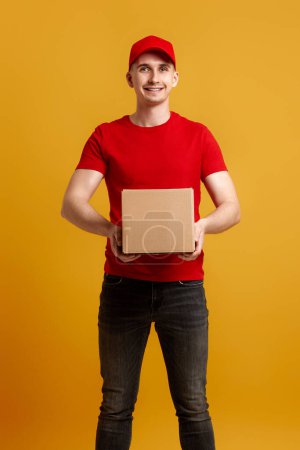 Téléchargez les photos : Livraison employé en bonnet rouge, t-shirt rouge tenant boîte en carton isolé sur fond jaune - en image libre de droit