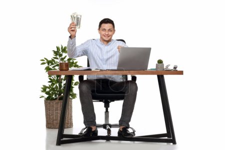 Téléchargez les photos : Jeune homme indépendant travaillant sur ordinateur portable et tenant de l'argent tout en étant assis sur la chaise au bureau. - en image libre de droit