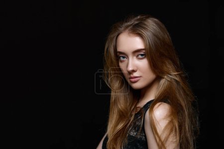 Téléchargez les photos : Magnifique femme brune aux cheveux longs en robe noire regardant la caméra sur fond noir. espace de copie - en image libre de droit