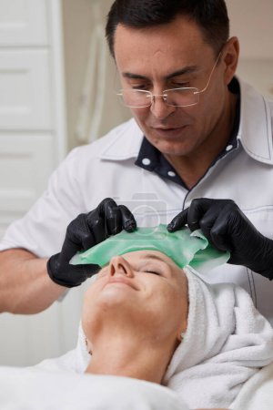Photo for Male beautician removing green alginate mask from face of beautiful woman in the spa salon. spa treatments - Royalty Free Image