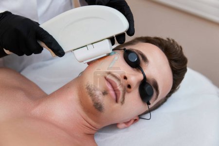 Photo for Young man receiving epilation in beauty center. Professional beautician doing laser hair removal procedure on male face. - Royalty Free Image