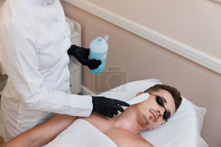 Foto de Esteticista aplica gel de enfriamiento a la cara masculina para recibir depilación en el centro de belleza. - Imagen libre de derechos