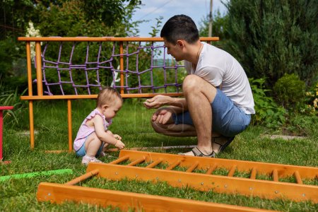 Photo for Dad with little child girl daughter collects wooden home sports complex outdoor in summer day - Royalty Free Image