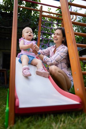 Photo for Happy little baby girl with mother playing on slide in playground in summer. Summer family leisure. - Royalty Free Image