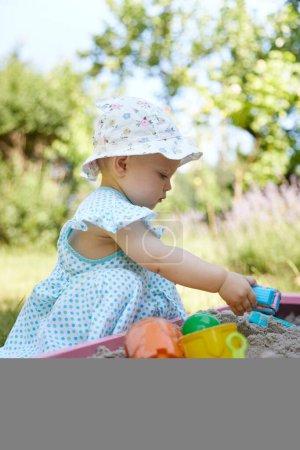 Foto de Niña linda jugando en arena en caja de arena con varios juguetes en el patio al aire libre en el día de verano soleado. - Imagen libre de derechos
