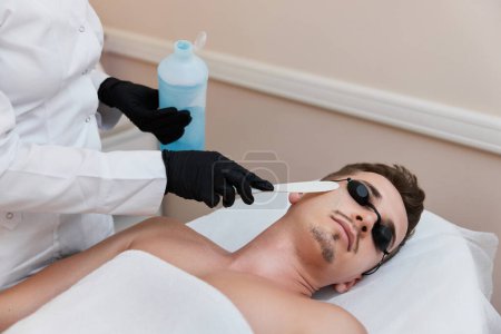 Photo for Beautician applies cooling gel to male facefor receiving epilation in beauty center. - Royalty Free Image