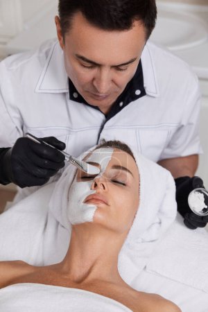 Foto de Esteticista masculino aplica la máscara a la cara de la hermosa mujer en el salón de spa. cuidado facial de la piel concepto. - Imagen libre de derechos