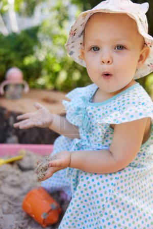 Foto de Feliz niña linda emocionada jugando en arena en caja de arena con varios juguetes en el patio al aire libre en el día de verano soleado. - Imagen libre de derechos