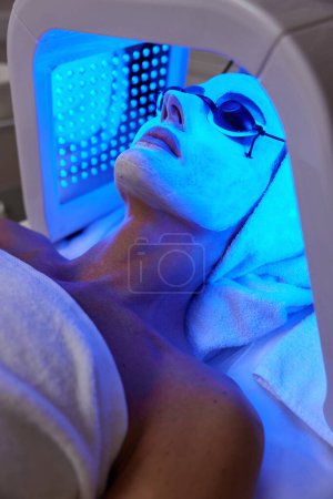 Frau mit Schutzbrille bekommt Gesichtsbehandlung mit LED-Therapie. LED-Maske in der Schönheitsklinik