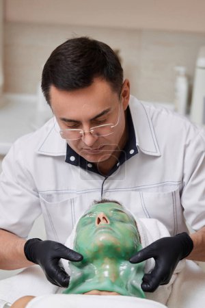 Téléchargez les photos : Esthéticienne masculine enlevant le masque d'alginate vert du visage de la belle femme dans le salon de spa. traitements spa - en image libre de droit