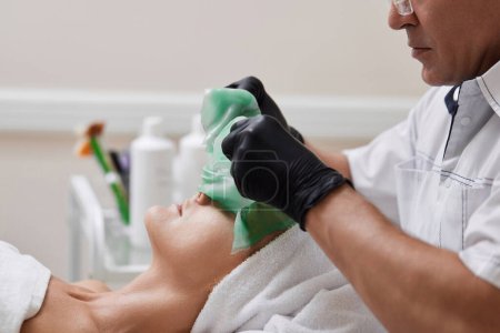 Téléchargez les photos : Esthéticienne masculine enlevant le masque d'alginate vert du visage de la belle femme dans le salon de spa. traitements spa - en image libre de droit