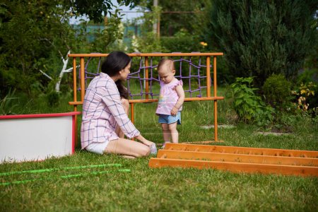 Photo for Mother with little child girl daughter collects wooden home sports complex outdoor. assembly process - Royalty Free Image