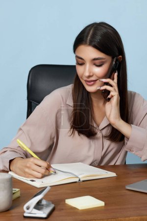 Téléchargez les photos : Belle femme d'affaires en chemise beige travaillant avec un ordinateur portable et ayant une conversation téléphonique avec le client - en image libre de droit