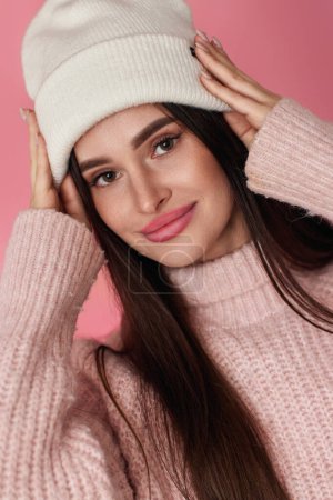 Photo for Portrait of happy playful woman keeps hands on white knitted hat on pastel pink background. close-up - Royalty Free Image