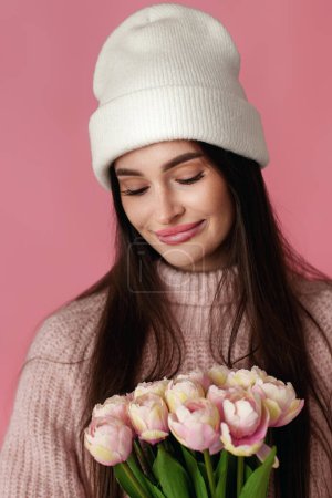 Téléchargez les photos : Jolie femme au chapeau blanc, pull tenant bouquet de tulipes sur fond rose pastel. - en image libre de droit