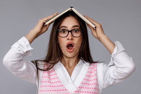 Téléchargez les photos : Femme brune surprise dans des lunettes avec livre sur la tête avec bouche ouverte sur fond gris - en image libre de droit