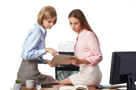 Photo for Two office female workers cooperating on project, sitting on desk with clipboard in office - Royalty Free Image