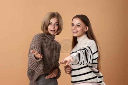 Téléchargez les photos : Deux amies ensemble dans des vêtements décontractés pointent index de côté sur fond beige. Femme blonde surprise et son amie - en image libre de droit