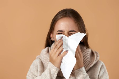 Photo for Unhappy young woman in sweater suffering from fever and flu, blowing nose in napkin on beige background - Royalty Free Image