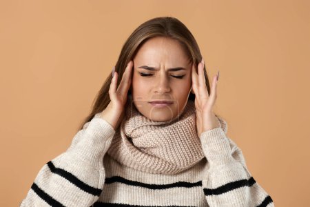 Téléchargez les photos : Jeune femme en pull en laine souffrant de maux de tête sur fond beige. douleur et migraine - en image libre de droit