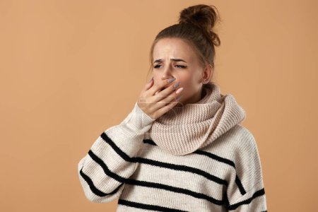 Téléchargez les photos : Malheureuse femme malade en pull toux couvrant la bouche avec la main sur fond beige. souffrant de grippe - en image libre de droit