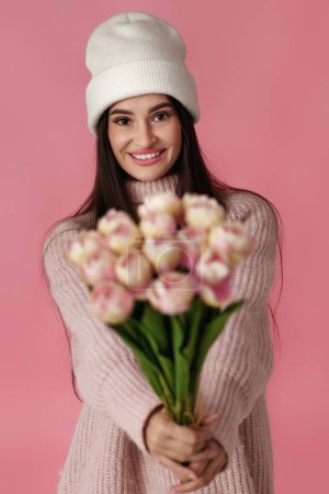 Photo for Pretty woman in white hat, sweater holding bouquet of tulips on pastel pink background. - Royalty Free Image