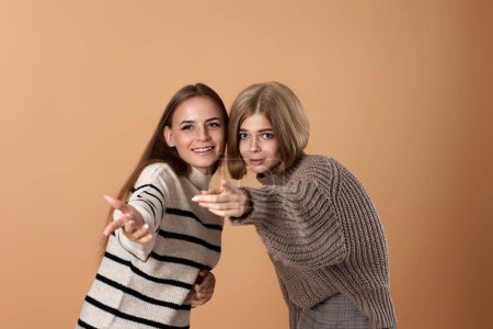 Foto de Dos amigas en ropa casual apuntan con el dedo índice a un lado sobre fondo beige. Mujer rubia sorprendida y su amiga - Imagen libre de derechos