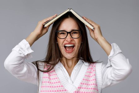 Photo for Surprised lovely brunette woman in glasses with book on head with open mouth on grey background - Royalty Free Image