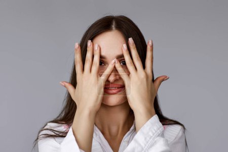 Foto de Bonita mujer cubriéndose los ojos con las manos y asomándose entre los dedos con expresión sorprendida sobre fondo gris. - Imagen libre de derechos