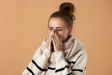Foto de Mujer joven infeliz en suéter que sufre de fiebre y gripe, sonando la nariz en servilleta sobre fondo beige - Imagen libre de derechos