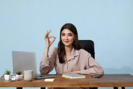 Téléchargez les photos : Dame heureuse montrant signe geste correct assis au bureau et en utilisant un ordinateur portable sur fond bleu - en image libre de droit