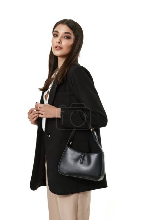 Téléchargez les photos : Belle femme avec sac à main en cuir noir en blazer noir et pantalon beige isolé sur fond blanc - en image libre de droit